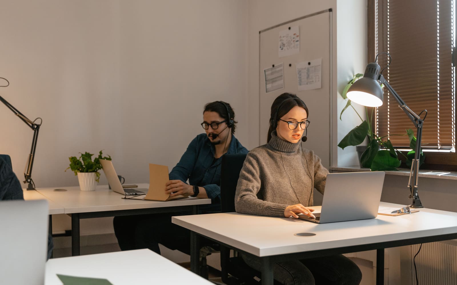 Two people wearing headsets while working at laptops in an office.